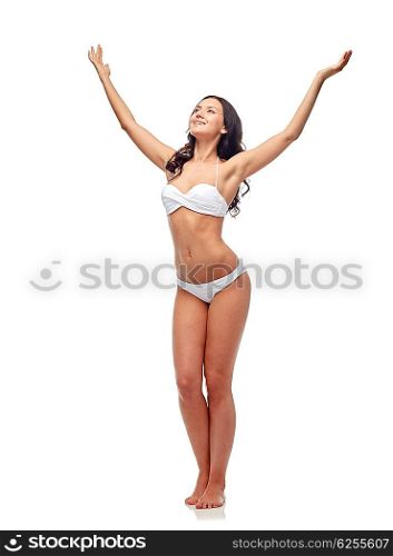 people, fashion, swimwear, summer and beach concept - happy young woman posing in white bikini swimsuit with raised hands