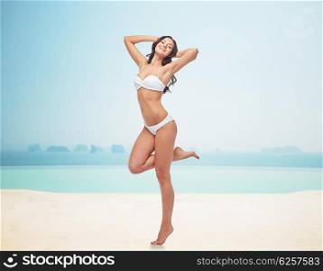people, fashion, swimwear, summer and beach concept - happy young woman posing in white bikini swimsuit with raised hands and standing on one leg over infinity pool at beach resort