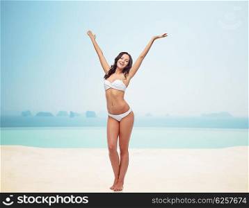 people, fashion, swimwear, summer and beach concept - happy young woman posing in white bikini swimsuit dancing with raised hands over infinity pool at beach resort