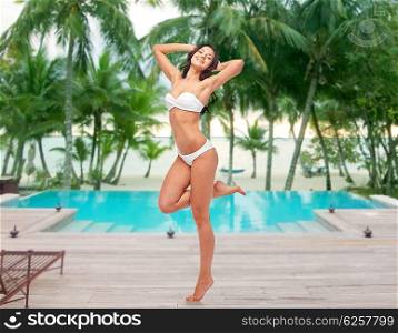 people, fashion, swimwear, summer and beach concept - happy young woman posing in white bikini swimsuit with raised hands and standing on one leg over swimming pool at beach resort