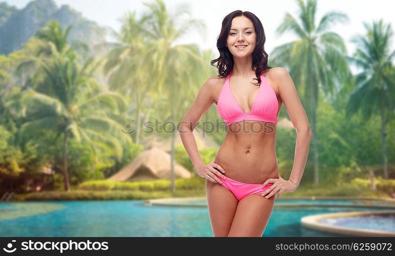 people, fashion, swimwear, summer and beach concept - happy young woman posing in pink bikini swimsuit over swimming pools and palm trees background