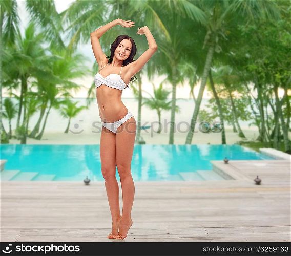 people, fashion, swimwear, summer and beach concept - happy young woman posing in white bikini swimsuit with raised hands over swimming pool at beach resort