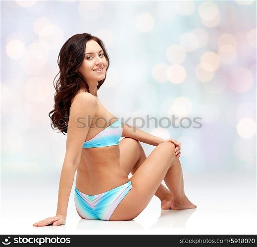 people, fashion, swimwear, summer and beach concept - happy young woman sunbathing in bikini swimsuit over blue holidays lights background