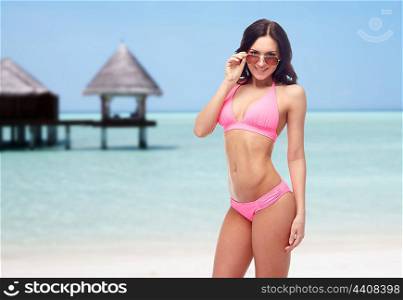 people, fashion, swimwear, summer and travel concept - happy young woman in sunglasses and pink swimsuit looking at you over maldives beach with bungalow background