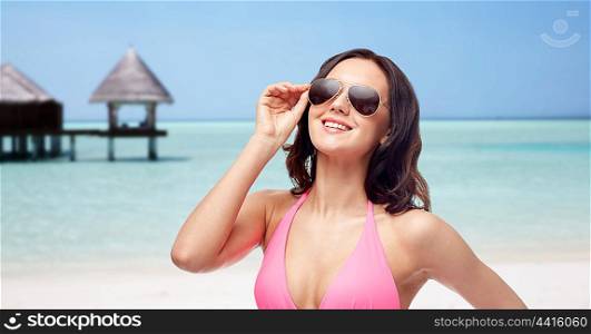 people, fashion, swimwear, summer and travel concept - happy young woman in sunglasses and pink bikini swimsuit over maldives beach with bungalow background