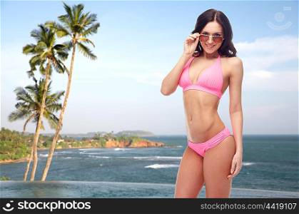 people, fashion, swimwear, summer and travel concept - happy young woman in sunglasses and pink swimsuit looking at you over Sri Lanka beach with palms swimming pool background