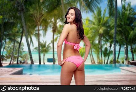 people, fashion, swimwear, summer and travel concept - happy young woman in pink bikini swimsuit looking back over tropical beach with swimming pool background