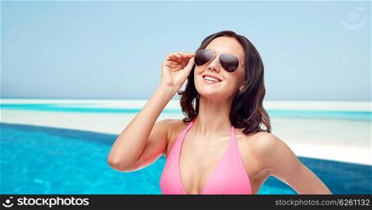 people, fashion, swimwear, summer and travel concept - happy young woman in sunglasses and pink bikini swimsuit over maldives beach with swimming pool background
