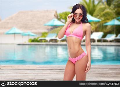 people, fashion, swimwear, summer and travel concept - happy young woman in sunglasses and pink swimsuit looking at you over resort swimming pool with parasols and sunbeds background