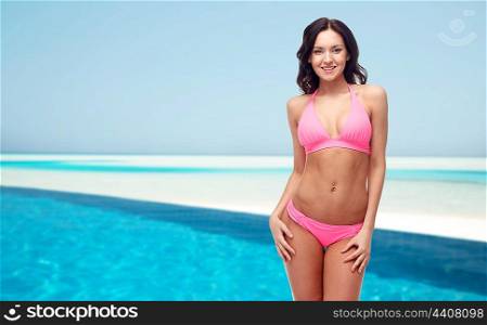 people, fashion, swimwear, summer and travel concept - happy young woman posing in pink bikini swimsuit over maldives beach with swimming pool background