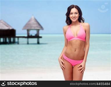 people, fashion, swimwear, summer and travel concept - happy young woman posing in pink bikini swimsuit over maldives beach with bungalow background
