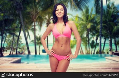 people, fashion, swimwear, summer and travel concept - happy young woman posing in pink bikini swimsuit over tropical beach with swimming pool background
