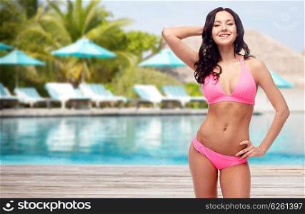 people, fashion, swimwear, summer and travel concept - happy young woman posing in pink bikini swimsuit over resort swimming pool with parasols and sunbeds background