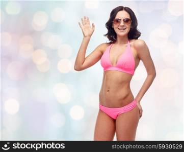 people, fashion, swimwear, summer beach and gesture concept - happy young woman in sunglasses and pink swimsuit waving hand over blue holidays lights background