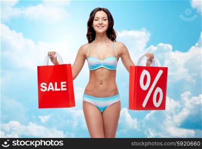 people, fashion, swimwear, summer sale and beach concept - happy young woman in bikini swimsuit with red shopping bags over blue sky and clouds background