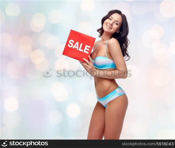 people, fashion, swimwear, summer shopping and beach concept - happy young woman in bikini swimsuit with red sale sign over blue holidays lights background