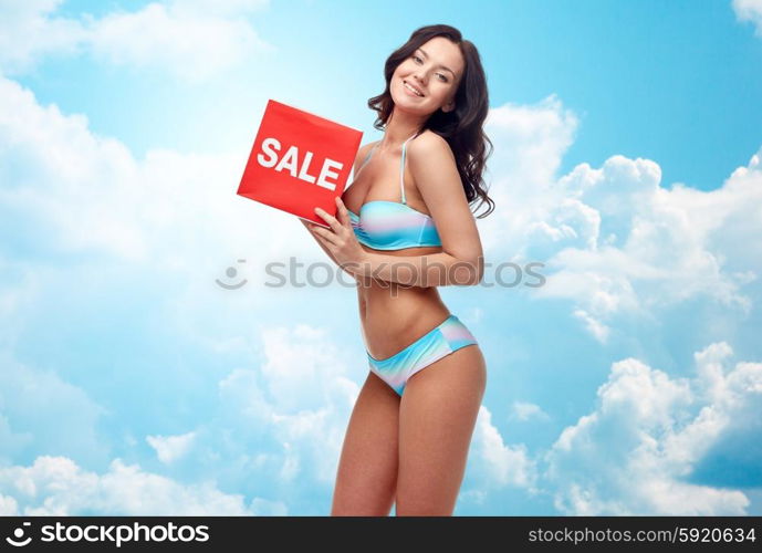 people, fashion, swimwear, summer shopping and beach concept - happy young woman in bikini swimsuit with red sale sign over blue sky and clouds background