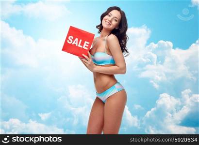 people, fashion, swimwear, summer shopping and beach concept - happy young woman in bikini swimsuit with red sale sign over blue sky and clouds background