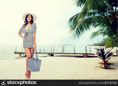 people, fashion, tourism, travel and summer concept - happy young woman in summer clothes and sun hat with bag over berth on tropical beach background