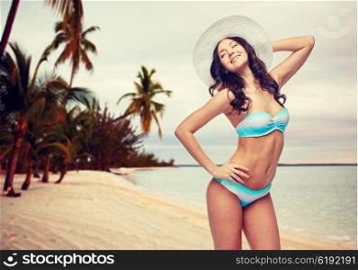 people, fashion, tourism, travel and summer concept - happy young woman in bikini swimsuit and sun hat posing over sunset on tropical beach background