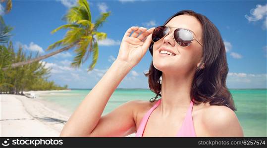 people, fashion, tourism, travel and summer concept - happy young woman in sunglasses and pink swimsuit looking up over tropical beach background