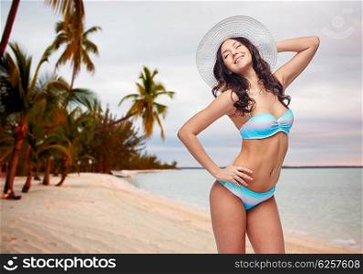 people, fashion, tourism, travel and summer concept - happy young woman in bikini swimsuit and sun hat posing over sunset on tropical beach background