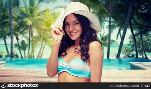 people, fashion, travel, tourism and summer concept - happy young woman in bikini swimsuit and sun hat over swimming pool and beach with palm trees background
