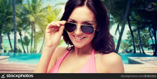 people, fashion, travel, tourism and summer concept - happy young woman in sunglasses looking at you over swimming pool and beach with palm trees background