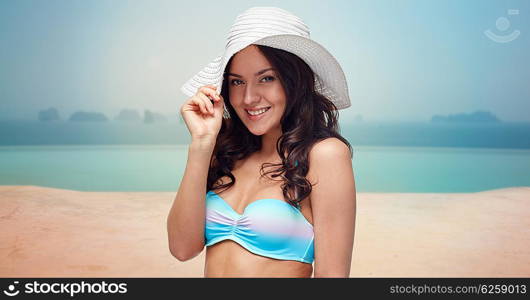 people, fashion, travel, tourism and summer concept - happy young woman in bikini swimsuit and sun hat over infinity pool at sea side background