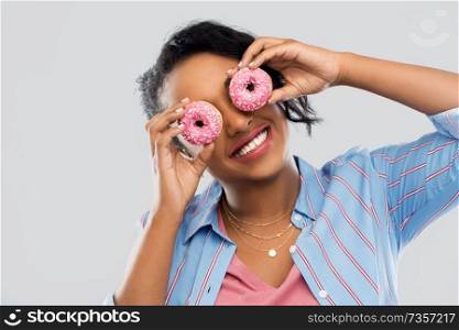 people, fast food and fun concept - happy african american young woman with donuts instead of eyes over grey background. happy african american woman with eyes of donuts