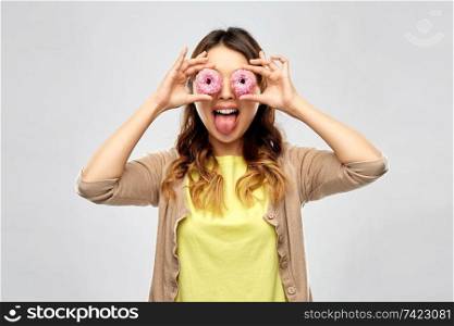 people, fast food and fun concept - happy asian young woman with donuts instead of eyes making faces over grey background. asian woman with eyes of donuts showing tongue