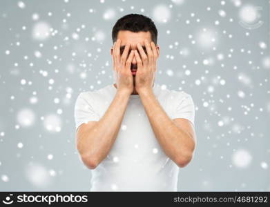 people, fear, emotions, winter and stress concept - man in white t-shirt covering his face with hands over snow on gray background