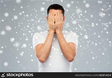 people, fear, emotions, winter and stress concept - man in white t-shirt covering his face with hands over snow on gray background