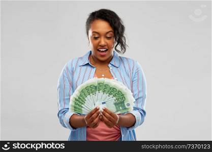 people, finances and wealth concept - surprised african american young woman holding fan of euro money over grey background. surprised african american woman with euro money