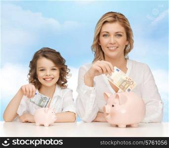 people, finances, family budget and savings concept - happy mother and daughter with piggy banks and paper money over blue sky background