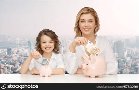 people, finances, family budget and savings concept - happy mother and daughter with piggy banks and paper money over city background