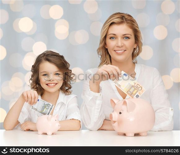 people, finances, family budget and savings concept - happy mother and daughter with piggy banks and paper money over lights background