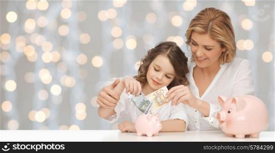 people, finances, family budget and savings concept - happy mother and daughter with piggy banks and paper money over lights background