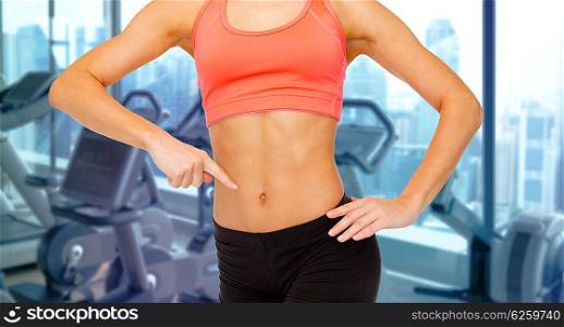 people, fitness and sport concept - close up of woman trainer pointing finger to her abdomen over gym machines background