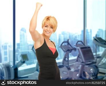 people, fitness, success, achievement and sport concept - happy woman fitness instructor with raised hand over gym machines background