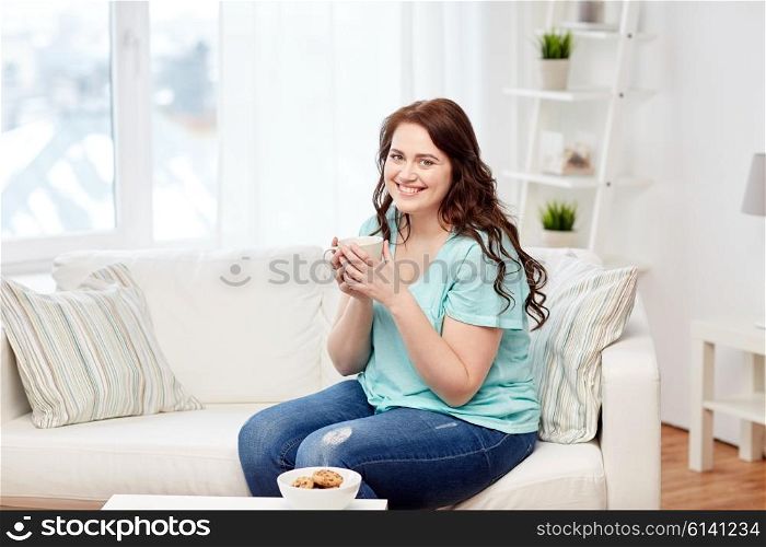 people, food, eating and leisure concept - happy plus size young woman with cup of tea with cookies at home
