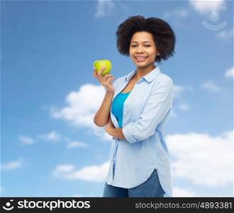 people, food, healthy eating and dental care concept - happy african american young woman with green apple over blue sky and clouds background