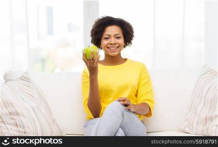 people, food, healthy eating and dental care concept - happy african american young woman with green apple sitting on sofa at home