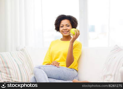 people, food, healthy eating and dental care concept - happy african american young woman with green apple sitting on sofa at home