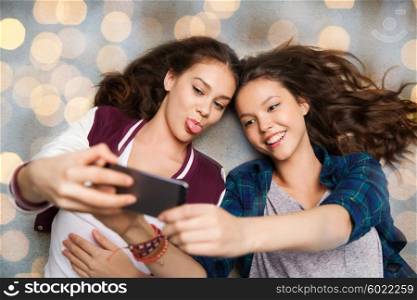 people, friends, teens and friendship concept - happy smiling pretty teenage girls lying on floor and taking selfie with smartphone over holidays lights background