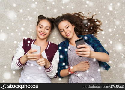 people, friends, winter, christmas and friendship concept - happy smiling pretty teenage girls lying on floor with smartphones over snow