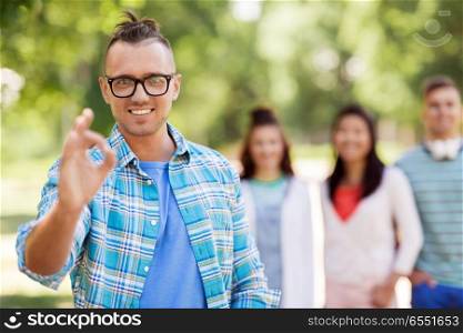 people, friendship and gesture concept - happy smiling man in glasses showing ok hand sign and group of happy friends outdoors. group of happy international friends outdoors. group of happy international friends outdoors