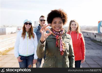 people, friendship and international concept - happy african american young woman or teenage girl in front of her friends showing ok sign on city street