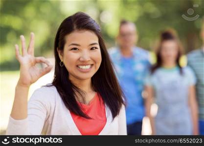 people, friendship and international concept - happy smiling asian woman showing ok hand sign and group of happy friends outdoors. group of happy international friends outdoors. group of happy international friends outdoors