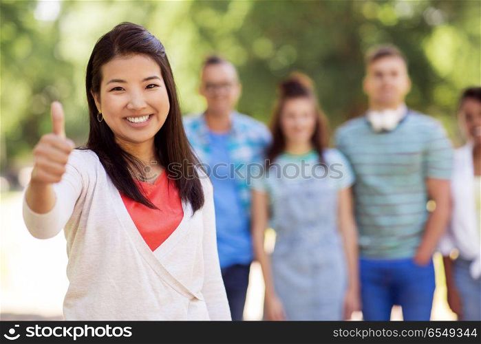 people, friendship and international concept - happy smiling asian woman showing thumbs up and group of happy friends outdoors. group of happy international friends outdoors. group of happy international friends outdoors